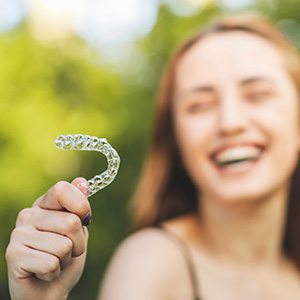 Woman holding Invisalign to foreground laughing with trees blurry in background